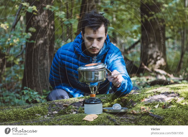 Man in a blue down jacket boils water for tea and food on a portable stove and in a stainless steel ultralight pot in the middle of the wilderness. Hiking lifestyle