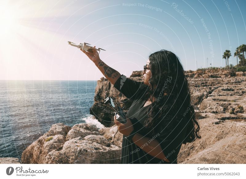 happy woman with drone, looking to the side black dress and remote control, on cliff, by the sea sunny day. technological concept adventure aerial air aircraft