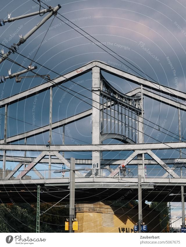 Arch and pillars of steel road bridge, cyclist on it, overhead wire in front of it, against dramatic sky Bridge Steel cyclists piers Overhead line Bicycle Sky