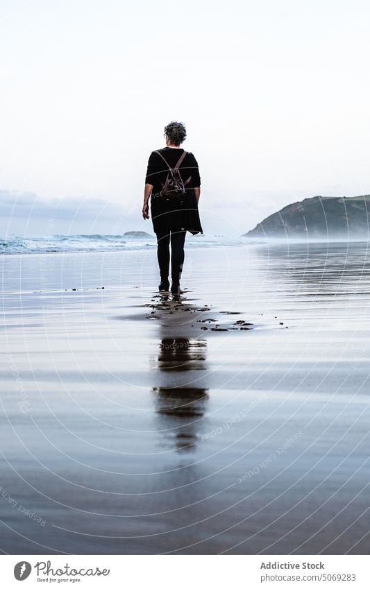 Senior woman walking on wet sand traveler sea beach resort weekend storm female elderly senior aged retire pensioner trip stroll holiday wave tourism tourist