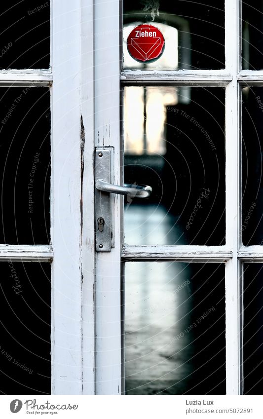 View through: an old white wooden door, behind it a dark corridor with window to the courtyard. Contrast to bw, light and dark only a bright red sticker with notice.