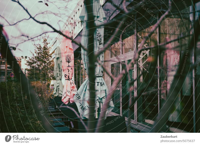 View through bare branches to the facade of an urban cafe, large glass panes in metal frame, folded umbrellas in front of it Looking Facade twigs Café Sunshade