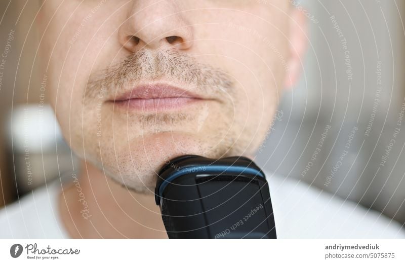 close up of caucasian man in white t shirt is shaving with electric razor at bathroom at home beard adult care handsome looking at camera machine male trimmer