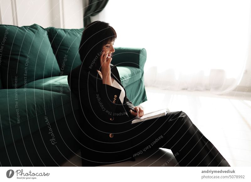 Businesswoman is talking on a smartphone. Tired brunette woman is sitting close to green sofa in a business black suit and high heels shoes with notebook. Green houseplant in interior. Work from home.