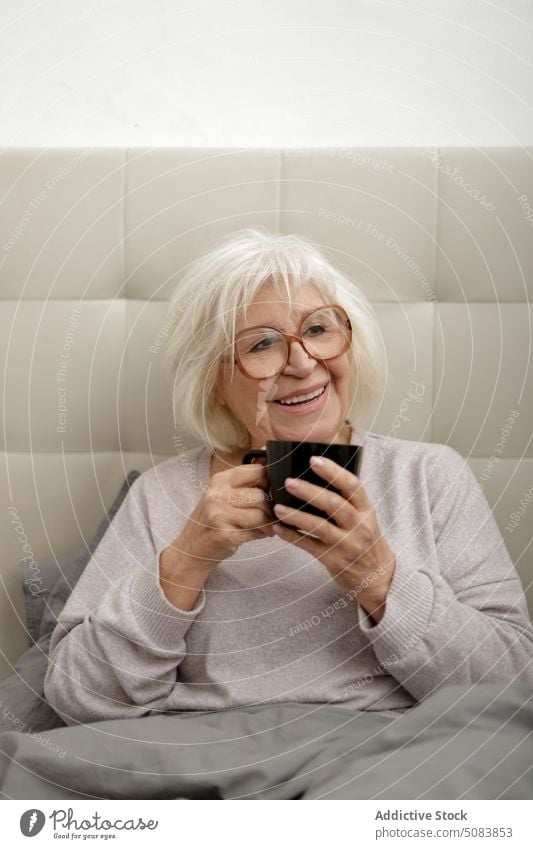 Positive senior woman drinking coffee in bed smile morning bedroom mug comfort beverage happy rest home cup female enjoy relax sweater at home apartment