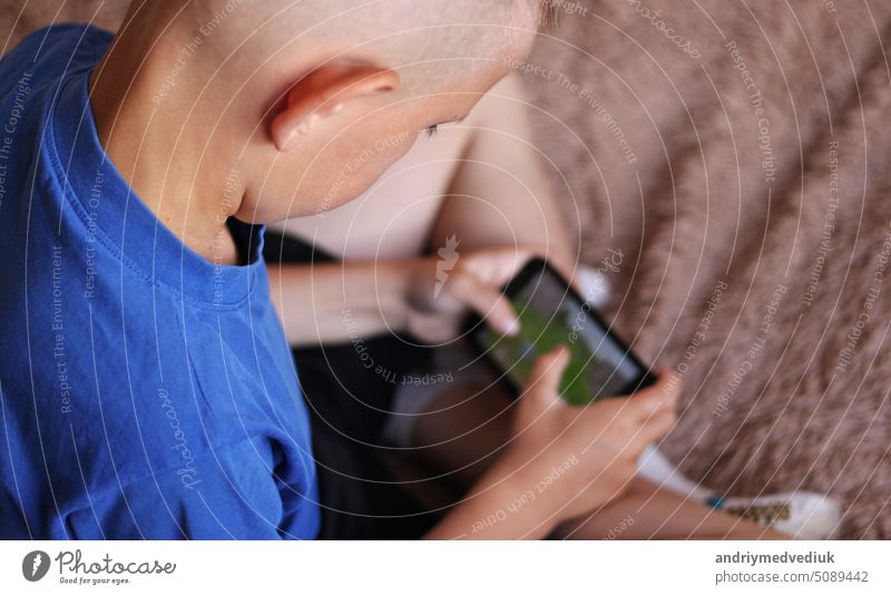 Preschool boy at Home is Lying on the bed, using a Smartphone. Child playing video games on a cellphone, Browses Internet, Views Content, Videos. Shooting wit camera over your shoulder