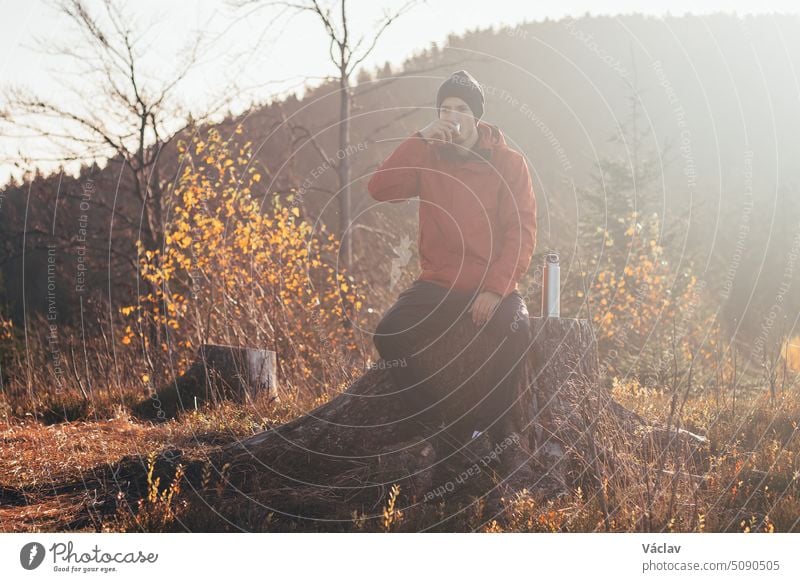 Tourist sitting on a fire pouring hot tea from a thermos into a cup at sunrise. A hiker is enlightened by the morning sun and enjoys the view. Beskydy mountains, Czech Republic