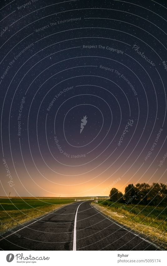 Night Starry Sky Above Country Road In Countryside And Green Field. Yellow City Lights On Background. Night View Of Natural Glowing Stars glowing evening field