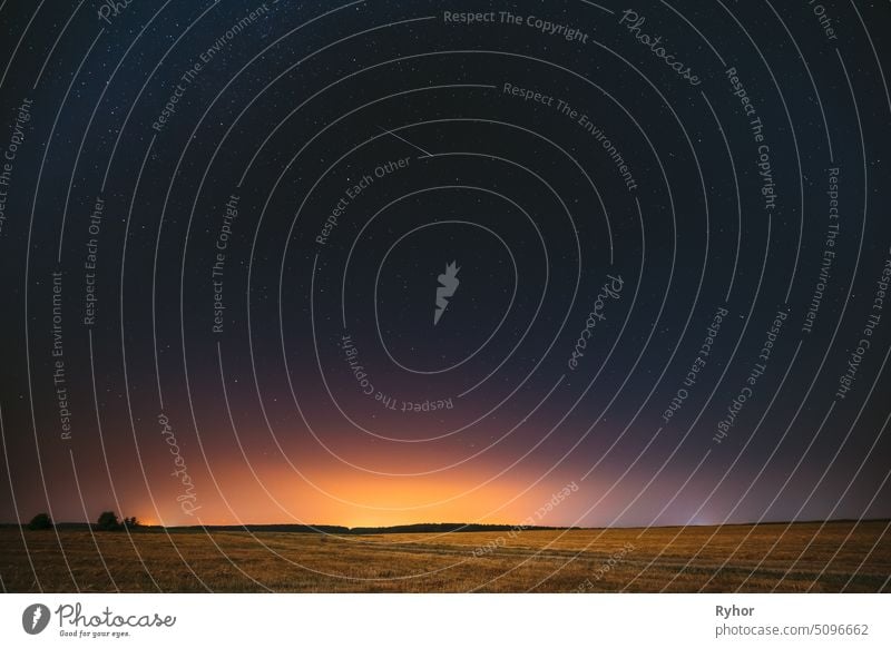 Natural Night Starry Sky Above Field Meadow. Glowing Stars, Meteorite Trail And Sunset Sunrise Lights Above Landscape In August Month. Meteoric Track In Night Sky Background