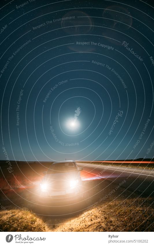 Night Starry Sky Above Country Asphalt Road In Countryside. Sedan Car Parking Near Asphalt Road In Summer Night. Yellow Car Lights And Night View Of Natural Glowing Stars