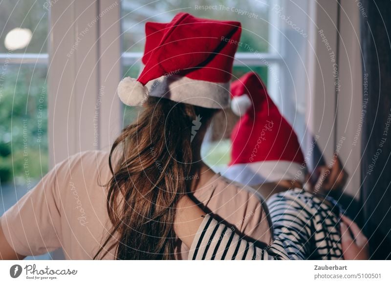 Two children with Christmas or Santa Claus caps look arm in arm out the window and wait for Christmas Child Wait mystery Joy Anticipation Red White Cap Infancy