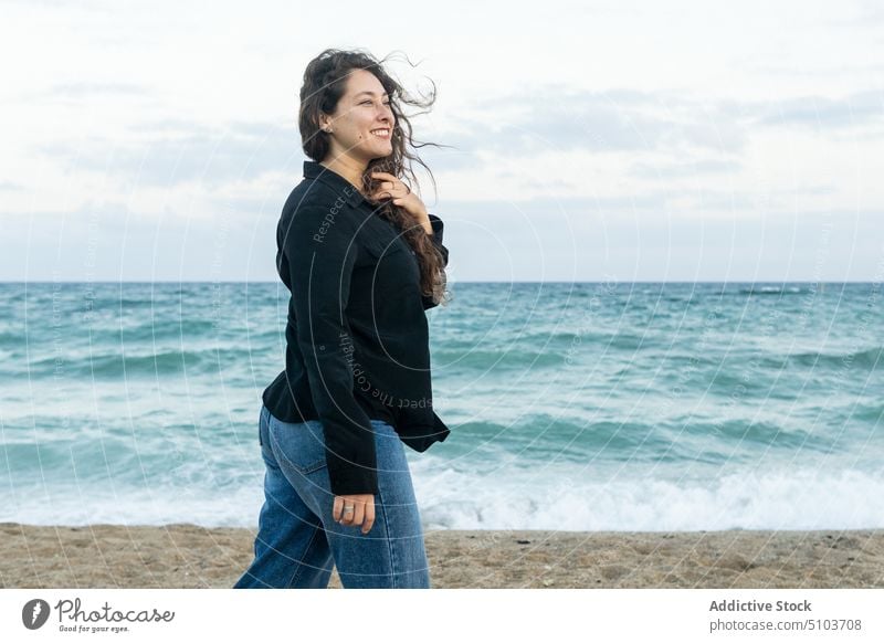 Happy curly haired woman standing on seashore beach sand water wave nature harmony seaside cloudy smile happy joy positive cheerful female young coast sky ocean