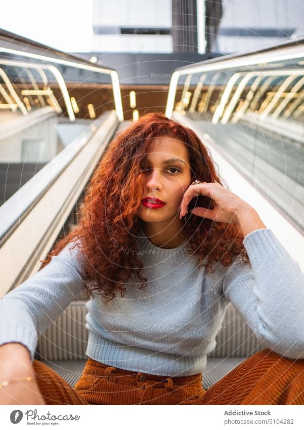 Stylish young woman sitting near escalator mall style appearance urban red hair curly hair touch face portrait female redhead sweater pants ginger personality