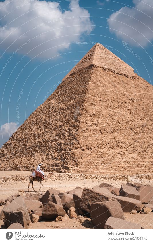 Anonymous local man riding camel near ancient pyramid in desert bedouin architecture ride sightseeing heritage travel explore sand historic tourism wonder