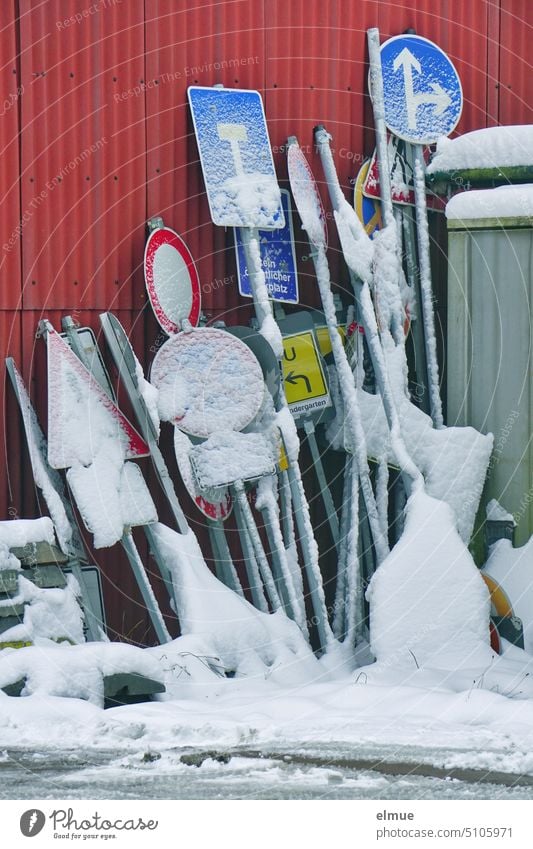 numerous traffic signs covered with snow leaning against red sheet metal wall / winter Road sign Collection Snow Winter Metal StVO Sign Signs Signs and labeling