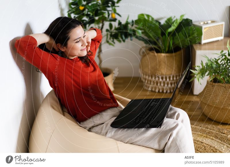 Relaxed young woman sitting in a bag chair using a laptop at home computer hassock couch happy leisure plants artificial relaxed relaxation smile wicker basquet