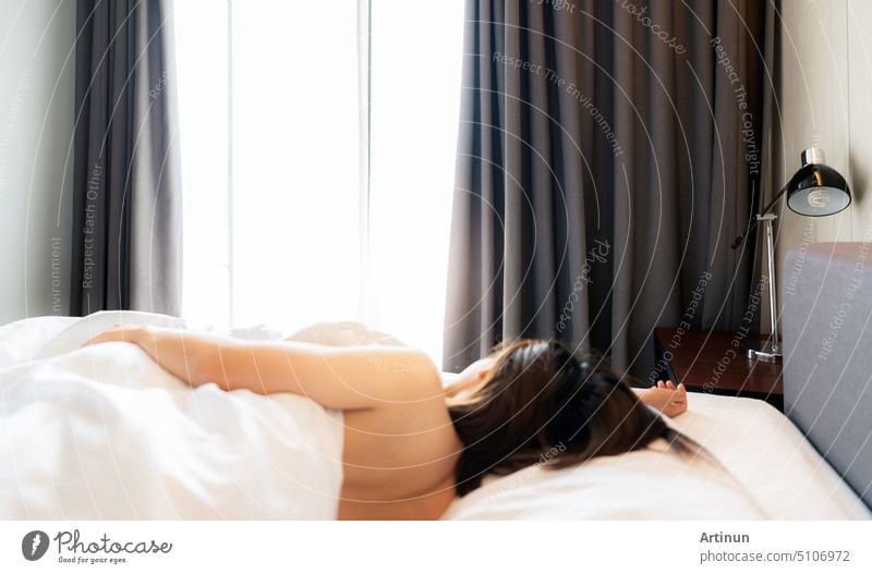 Side bed lamp and blur woman sleeping topless on bed in linen blanket at hotel or home bedroom. Back view of girl. Woman sleep on white comfort bed and duvet near window curtain and lamp. Lazy Sunday.