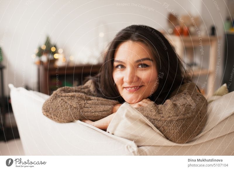 Beautiful caucasian woman smiling at her cozy home apartment leaning over the couch. Casual lifestyle female portrait. Millennial living. Expat life. Creating comfortable space around you.