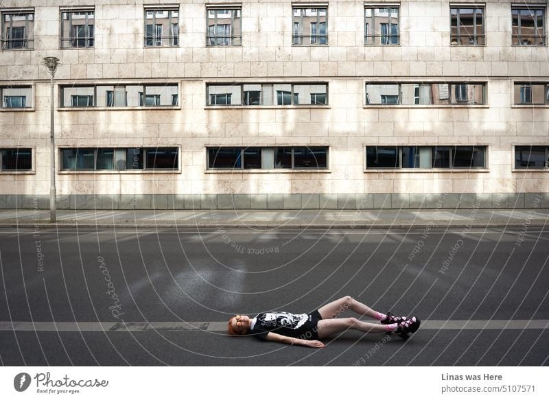 A fashionable street in Berlin where a red hair model is lying on the floor while being all gorgeous. Concrete surrounds her. Her high heels, pink socks, black clothes, and fancy sunglasses accompany her killer look.