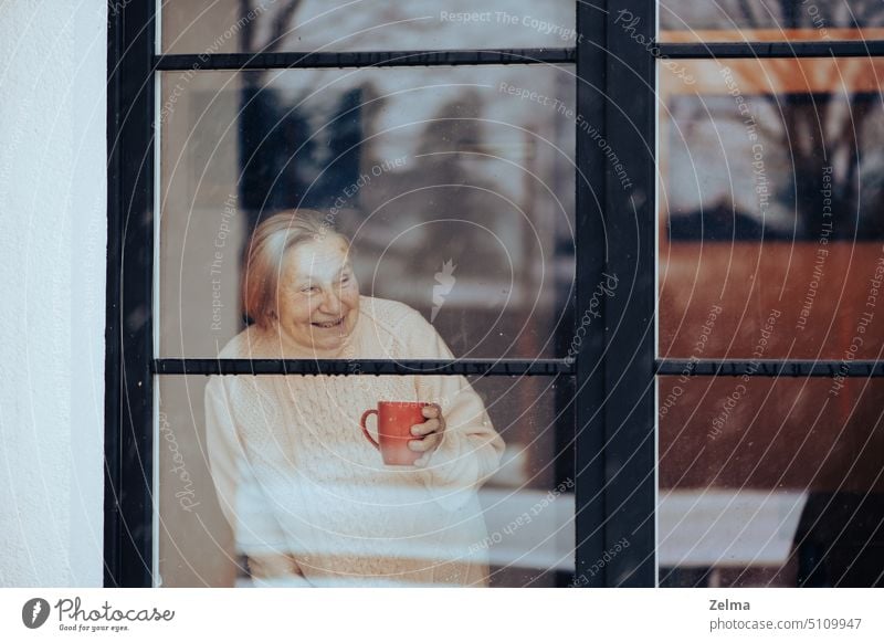 smiling old woman with a cup of coffee looks through a dirty window glass senior elderly female looking smile face nursing home house indoors loneliness sad