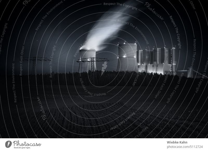 Power plant with long exposure at night. With empty field in the foreground and a forest with power poles in the background. Black and white photo with blue tinted sky. .