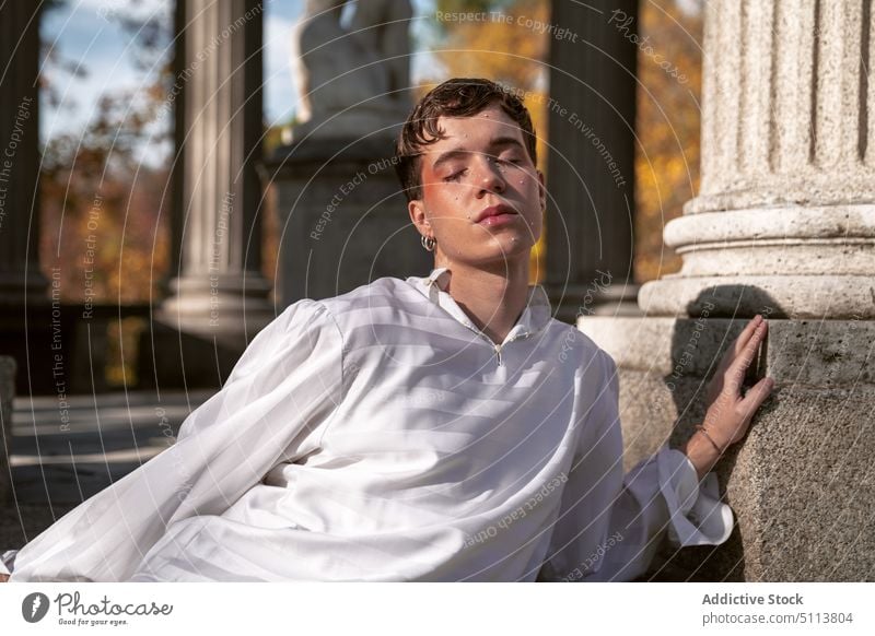 Calm homosexual man leaning on pillar in autumn park androgynous eyes closed lgbt dreamy column non binary portrait gender fluid pensive male young makeup