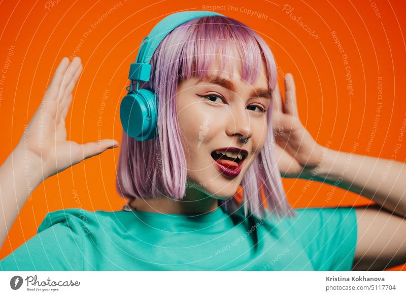 Exceptional woman with dyed violet hair listening music in headphones and singing on orange background. Unique hipster girl dancing person female fun studio