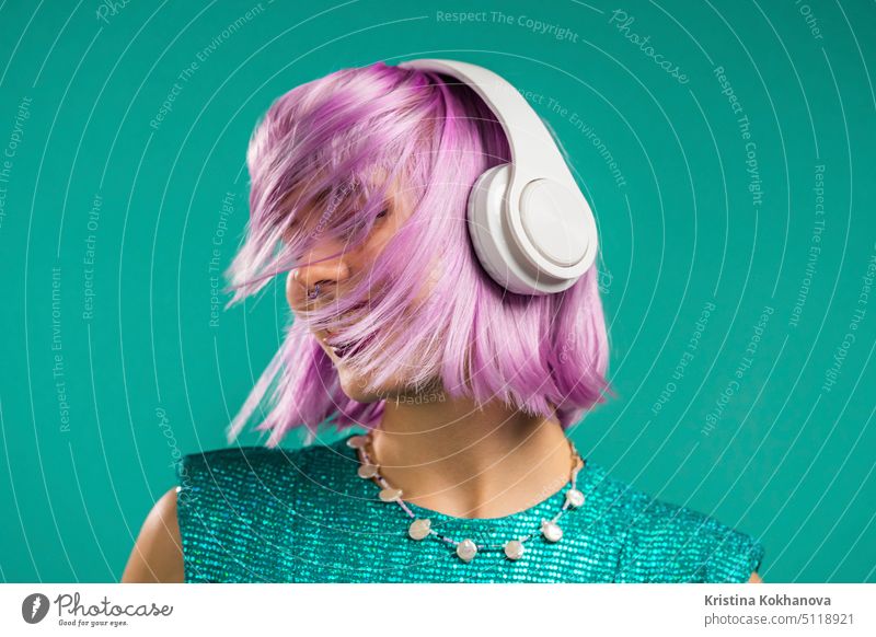 Unusual trendy woman with dyed violet hair listening music, dancing in headphones on turquoise background. Fashionista teen girl person female fun studio beauty