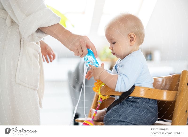 Happy infant sitting at dining table and playing with his toy in traditional scandinavian designer wooden high chair in modern bright atic home superwised by his mother