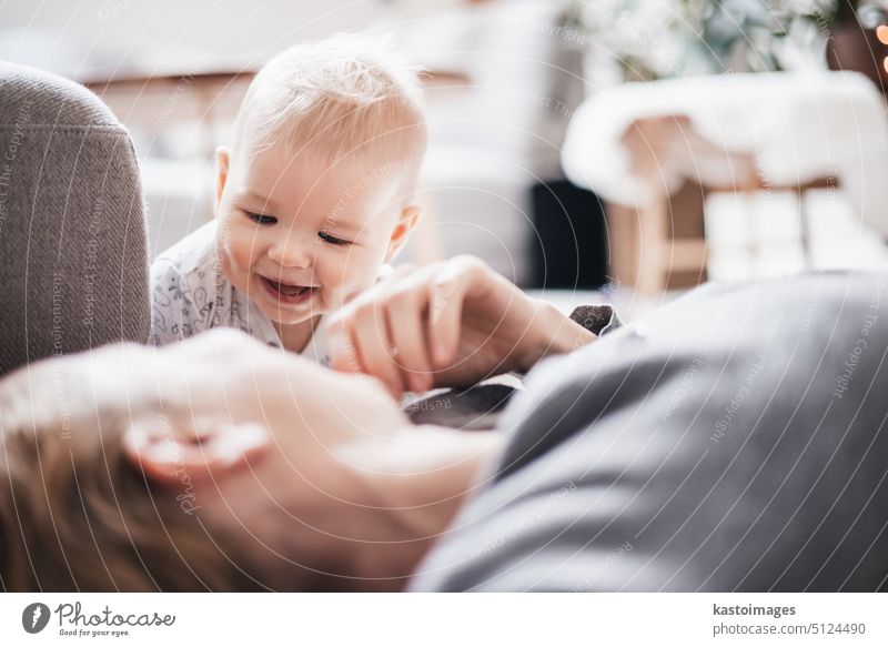 Happy family moments. Mother lying comfortably on children's mat playing with her baby boy watching and suppervising his first steps. Positive human emotions, feelings, joy.