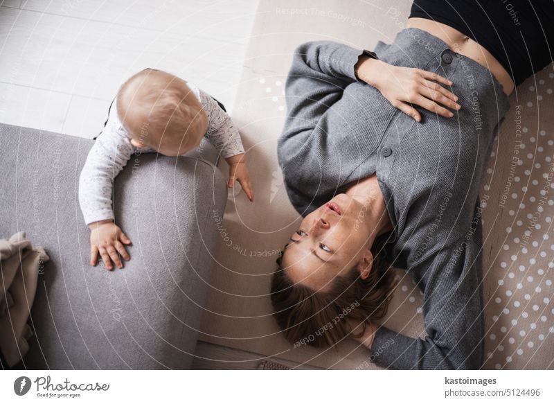 Happy family moments. Mother lying comfortably on children's mat playing with her baby boy watching and suppervising his first steps. Positive human emotions, feelings, joy.