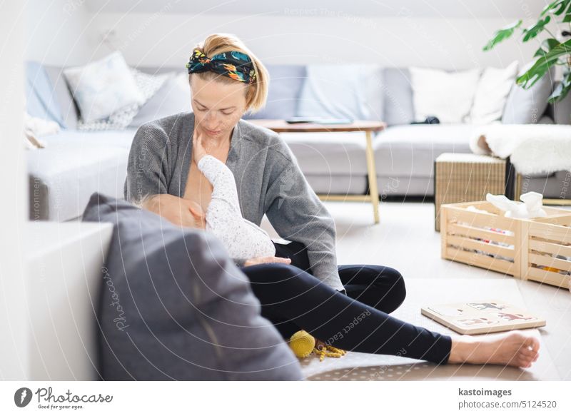 Young woman breastfeeding her infant baby boy casualy sitting on child's playing mat on living room floor at home. parent mother motherhood family milk kid care