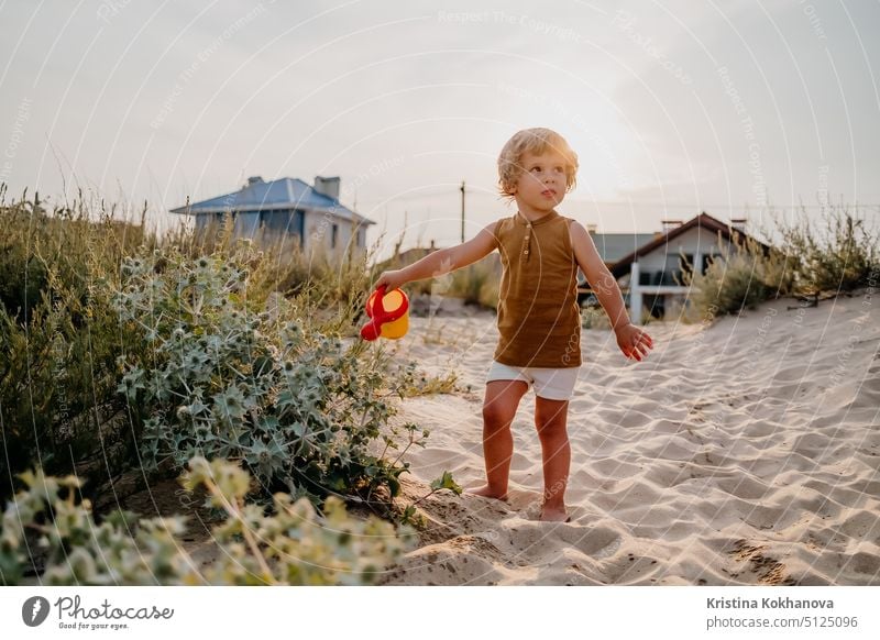 Cute little boy watering plants with can on sandy beach. summer sunny day. Toddler with colorful pot. Natural aestetic portrait of child. fun person vacation