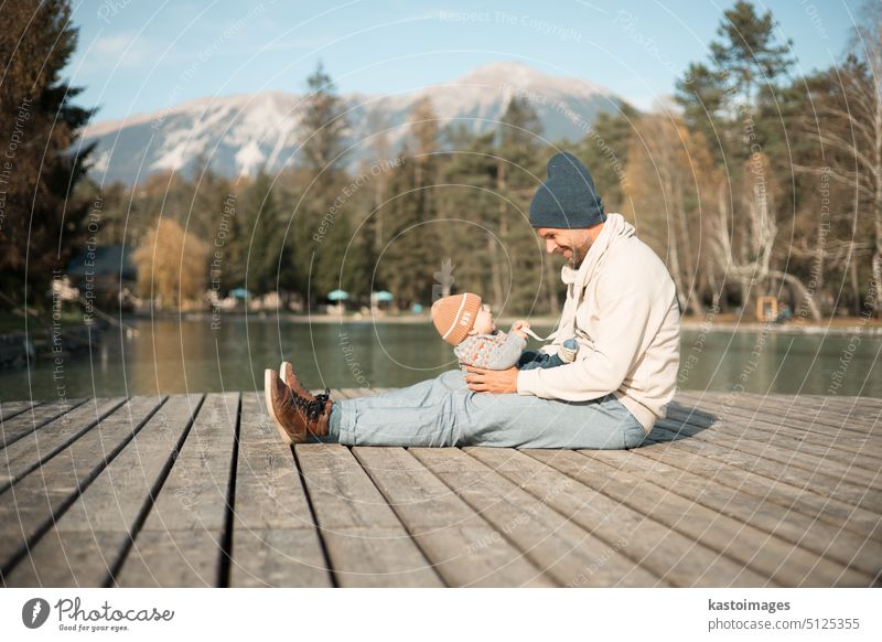 Happy family. Father playing with her baby boy infant oudoors on sunny autumn day. Portrait of dad and little son on wooden platform by lake. Positive human emotions, feelings, joy.