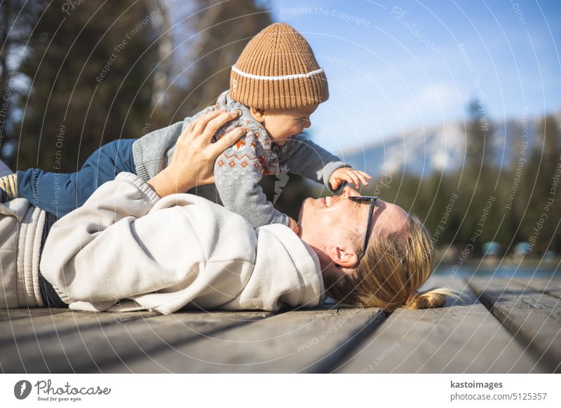 Happy family. Young mother playing with her baby boy infant oudoors on sunny autumn day. Portrait of mom and little son on wooden platform by lake. Positive human emotions, feelings, joy.