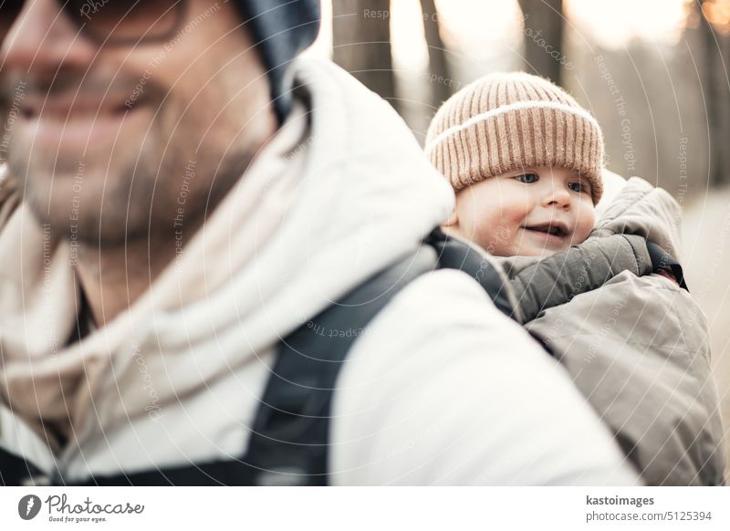 Sporty father carrying his infant son wearing winter jumpsuit and cap in backpack carrier hiking in autumn forest. travel parent wanderlust trekking together