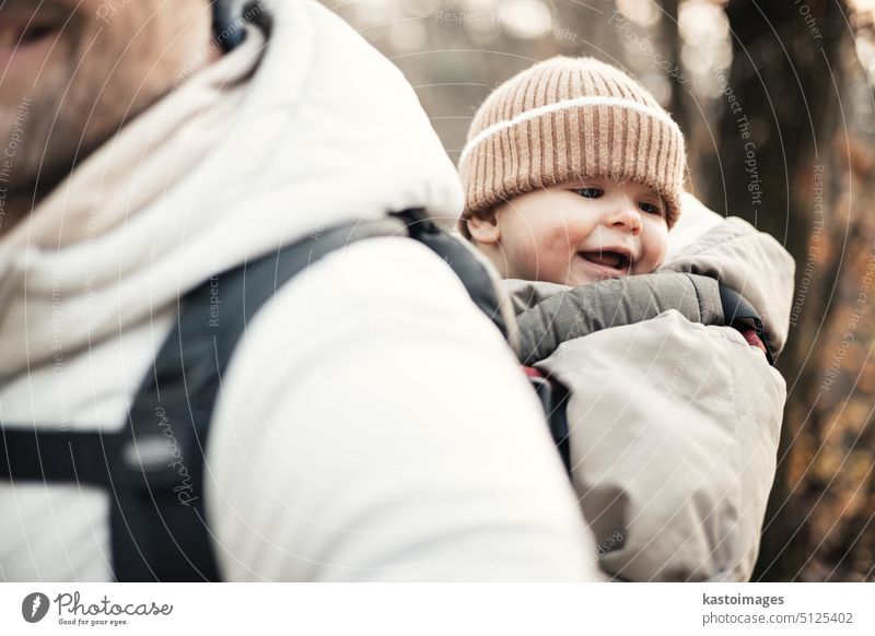 Sporty father carrying his infant son wearing winter jumpsuit and cap in backpack carrier hiking in autumn forest. travel parent wanderlust trekking together