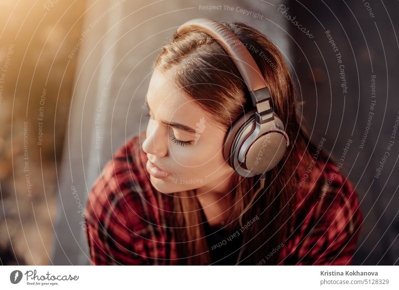 Young teenager listens to music through headphones in park.Girl in red plaid shirt smiles, dancing to rhythm.Concept of student life, freedom, modern youth