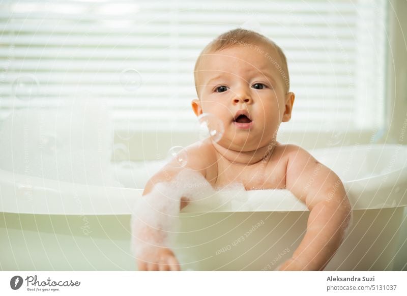 Cute little baby sitting in white bathtub with foam and soap bubbles. Taking bath and playing with toys. Baby hygiene. 6 month 6 month baby adorable bathe