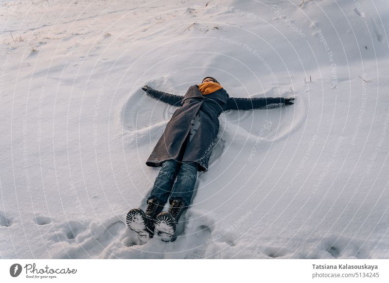 A woman in a coat, scarf and boots lies in a snow-covered field and makes a snow angel winter female warm clothes people person European White adult mid adult