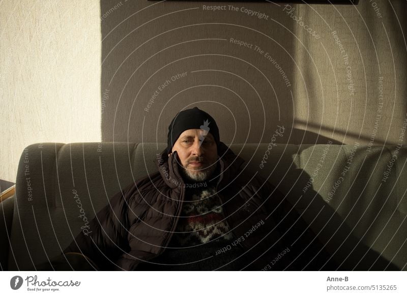 a man sits in thick winter clothes on a sofa in a room, hotel room, apparently waiting. He does not look at the viewer. Man Man with beard portrait Accidental