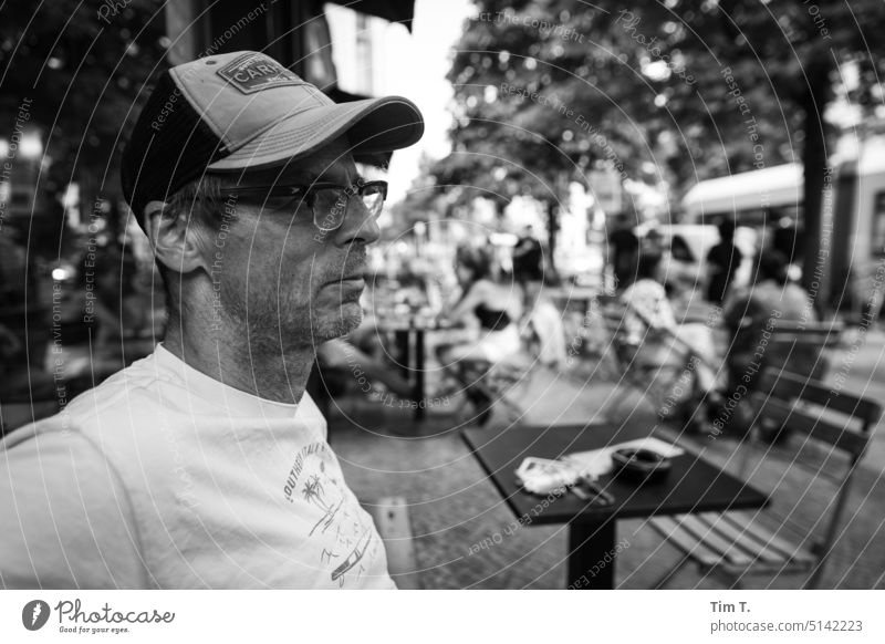 A man with peaked cap and glasses in front of a cafe in Berlin Prenzlauer Berg Man Eyeglasses Café chestnut avenue Exterior shot Town Downtown Old town