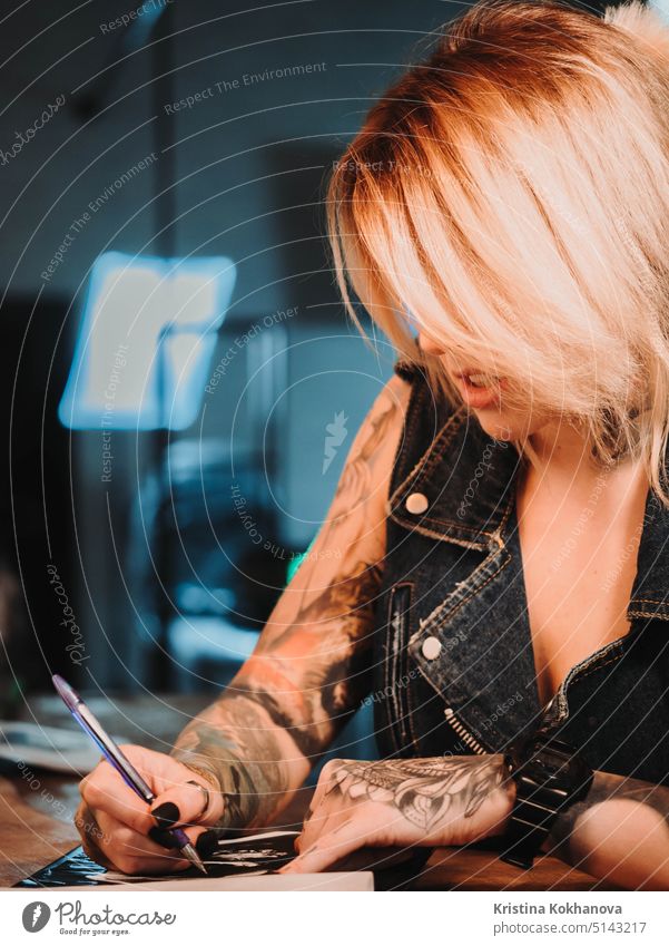 Tattoo artist drawing illustration of skull inside ink studio. Woman with black nails and rings at work. New fashion lifestyle artistic trends concept . Warm cinematic filter.