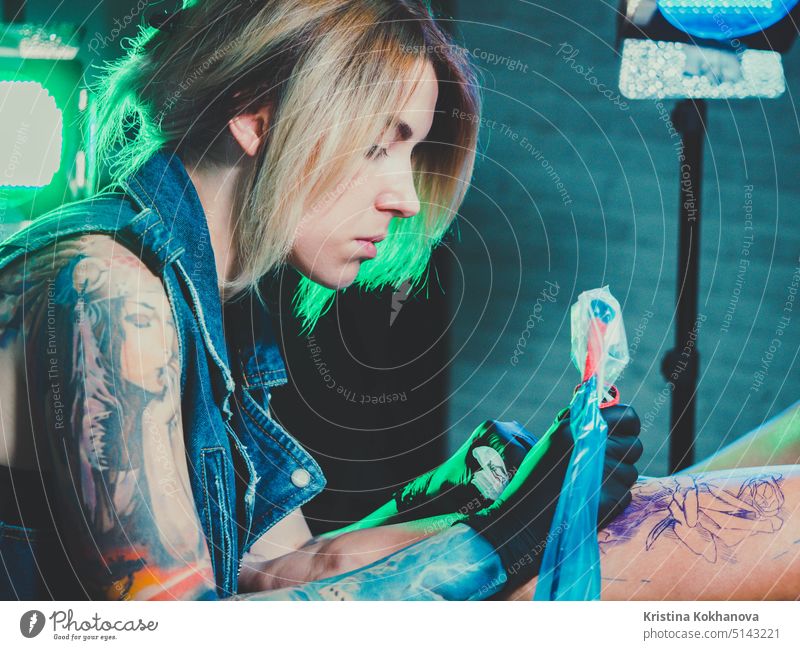 View of professional blonde tattoo female master is painting with black ink, making cover. Works in sterile latex gloves with handmade rotor gun machine in studio.