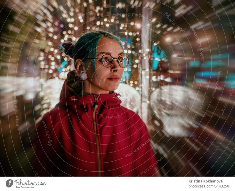 Young beautiful girl with blue dyed unusual hair, transparent glasses, piercing standing at night street with neon lights background. Portrait of happy cute stylish teenager.