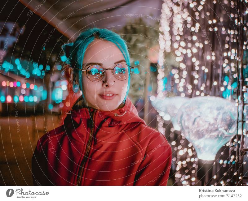 Young beautiful girl with blue dyed unusual hair, transparent glasses, piercing standing at night street with neon lights background. Portrait of happy cute stylish teenager.