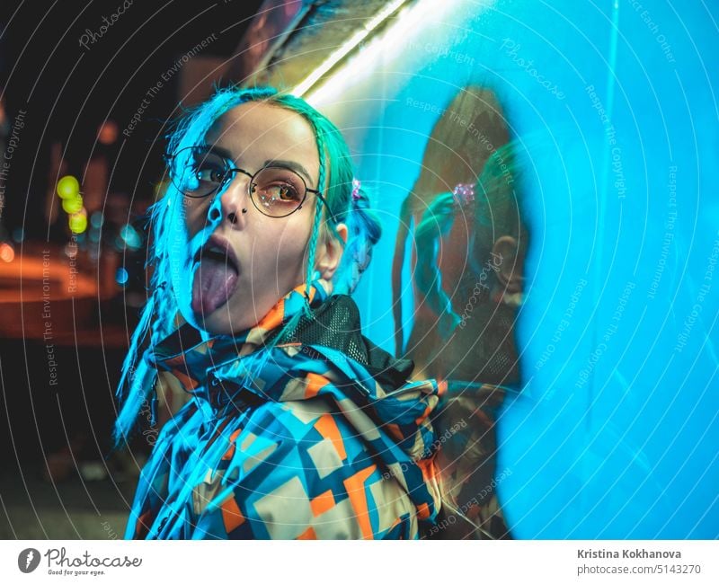 Young pretty girl with unusual hairstyle showing tongue near glowing turquoise neon light of the city at night. Dyed blue hair in braids. Pensive hipster teenager in glasses.