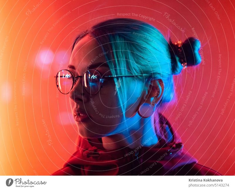 Millennial pretty girl with unusual hairstyle near glowing red neon of city at night. Dyed blue hair in braids. Mysterious hipster teenager in glasses. Reflection of light.