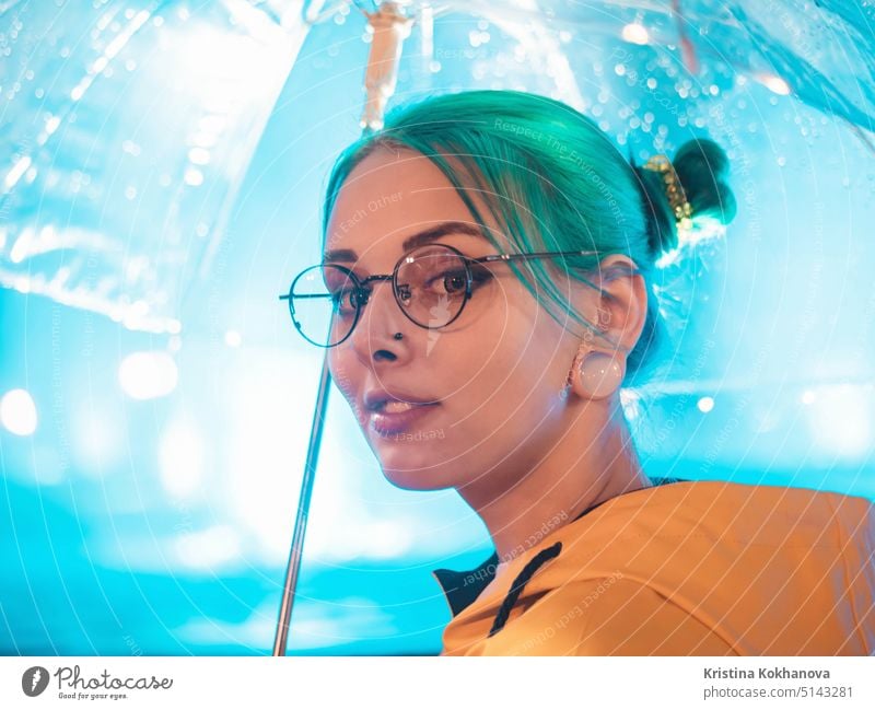 Young pretty girl with blue dyed hair in yellow raincoat and with transparent umbrella stands near fountain. Night neon illumination of city. Portrait of stylish hipster with glasses.