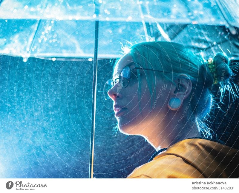 Young pretty girl with blue dyed hair in yellow raincoat and with transparent umbrella stands near fountain. Night neon illumination of city. Portrait of stylish hipster with glasses.